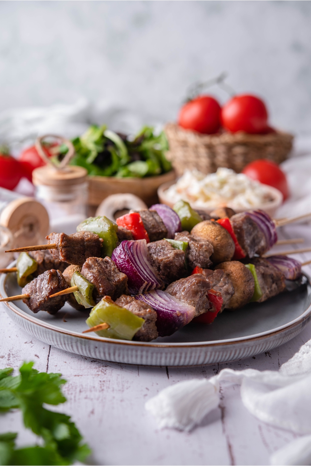 A stack of five baked steak kebabs filled with seasoned steak, bell peppers, mushrooms, and red onions. In the background are bowls of coleslaw, salad, tomatoes, and spices.