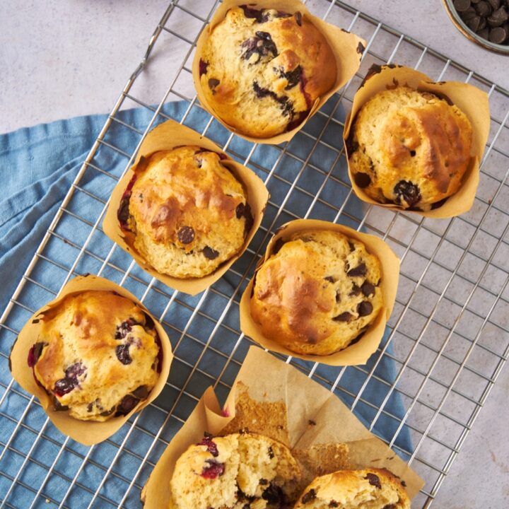Six freshly baked chocolate chip and blueberry muffins on a wire rack with one muffin cut in half.