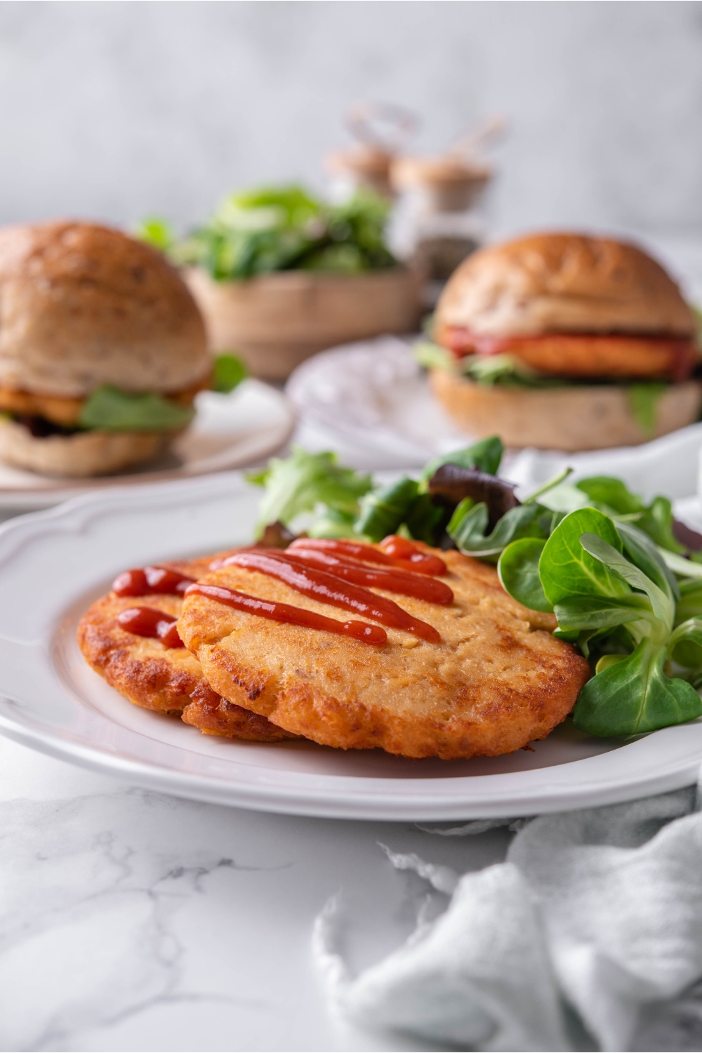 Two fried salmon patties covered with a drizzle of ketchup and layered on top of each other on a white plate with a side salad. In the background are two salmon burgers.