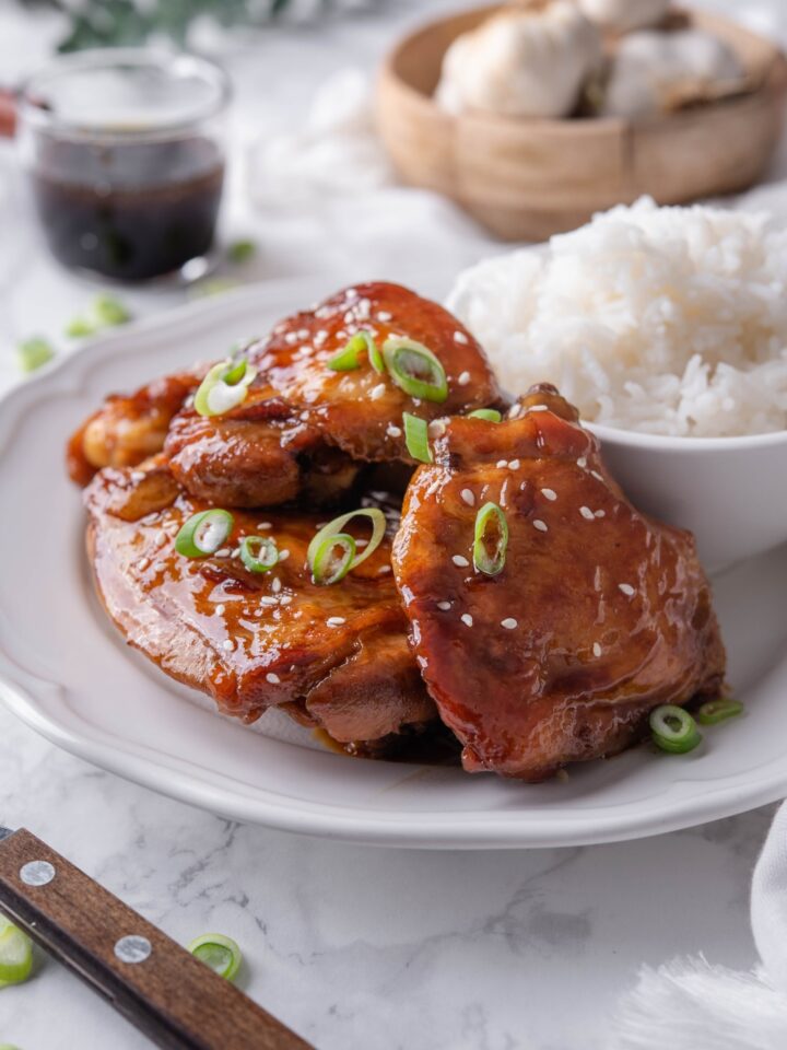 A plate of shoyu chicken thighs piled on top of each other with a side of white rice. The chicken is garnished with diced green onions and sesame seeds.