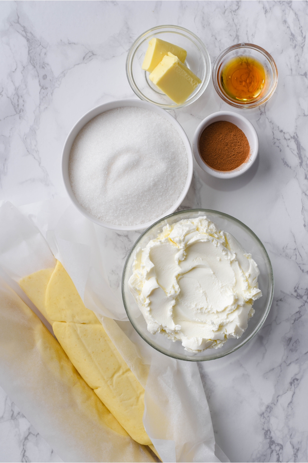 An assortment of ingredients including bowls of cream cheese, white sugar, vanilla extract, cinnamon, butter, and a roll of crescent dough wrapped in parchment paper.