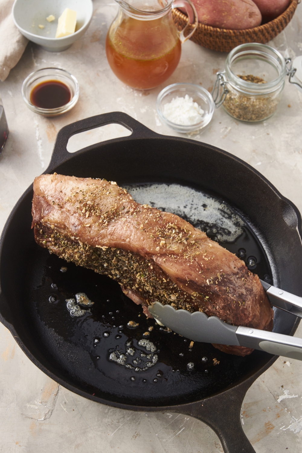 A prime rib being seared on one side in a cast iron skillet with a pair of tongs holding the steak upright.