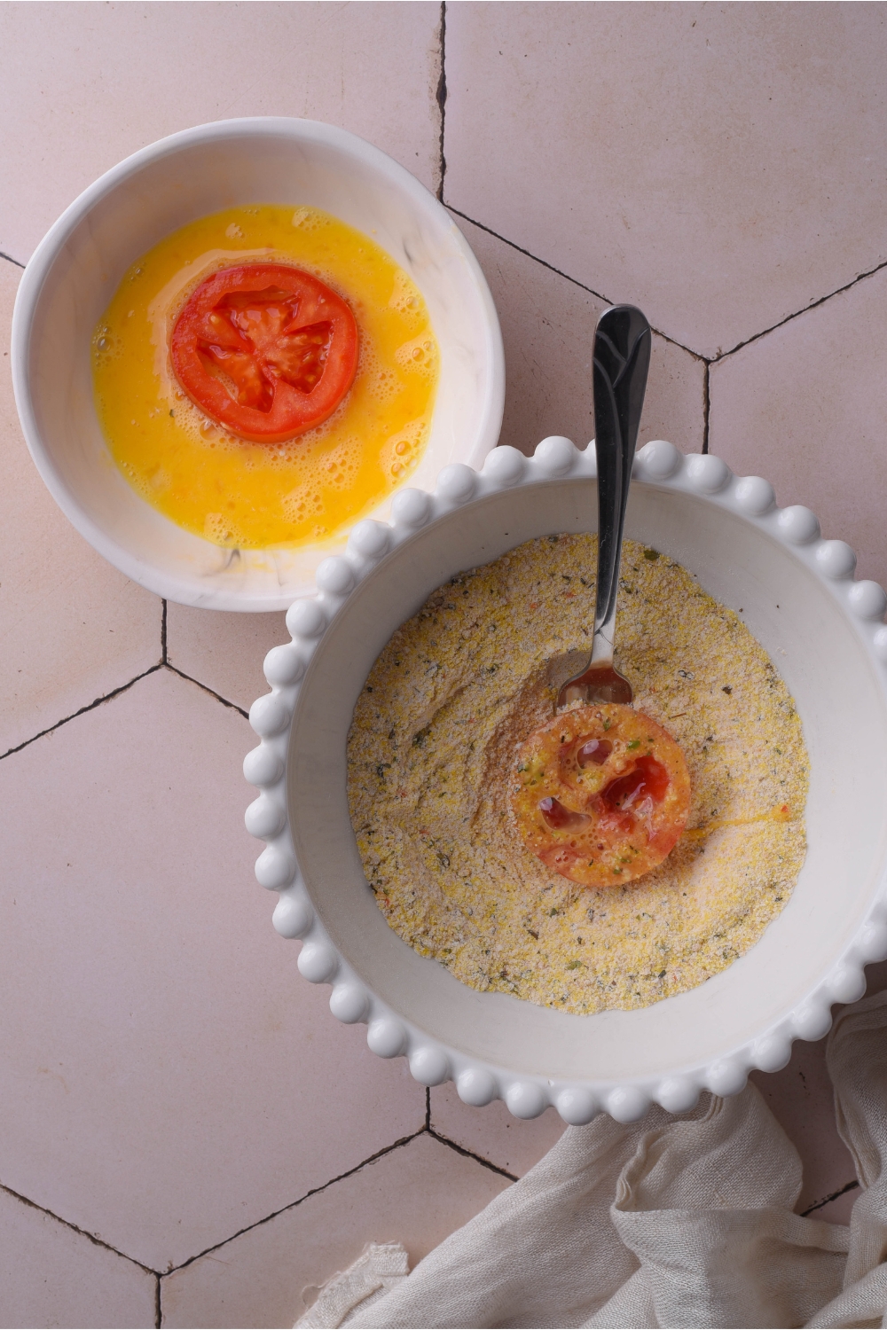 Two white bowls, one filled with an egg wash with a tomato slice in it and another filled with a cornmeal and bread crumb mixture with a spoon and a tomato slice.