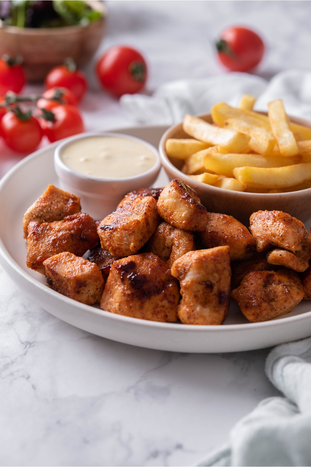 A pile of grilled chicken nuggets on a white plate with a side of french fries and a bowl of dipping sauce.