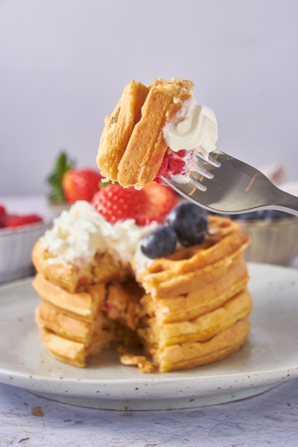 A forkful of waffles with whipped cream and in the background is the stack of waffles with a bite taken out.