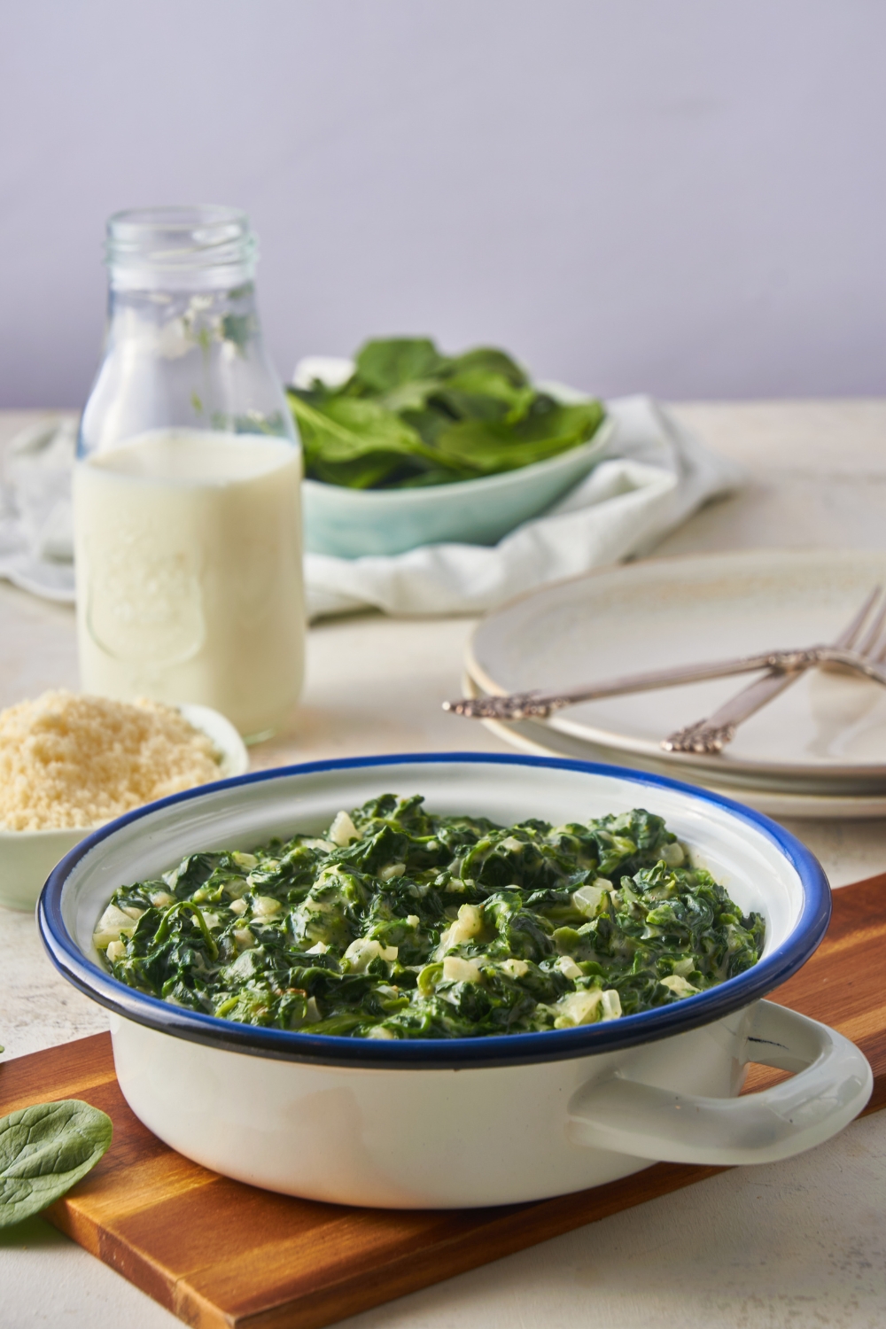 A white and blue rimmed serving dish filled with creamed spinach. The dish is atop a wooden board and there is a jar of milk in the background.