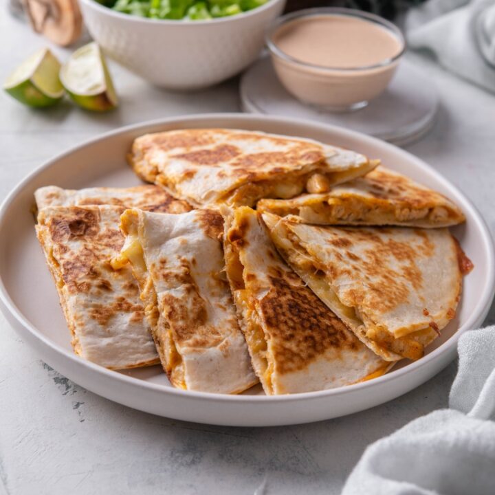 A plate of chicken quesadilla wedges piled on top of each other next to a bowl of quesadilla sauce and a bowl of lettuce.