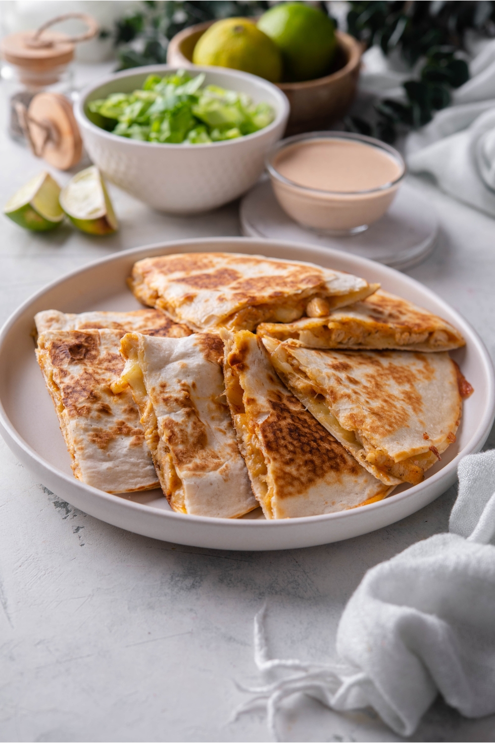 A plate of chicken quesadilla wedges piled on top of each other next to a bowl of quesadilla sauce and a bowl of lettuce.
