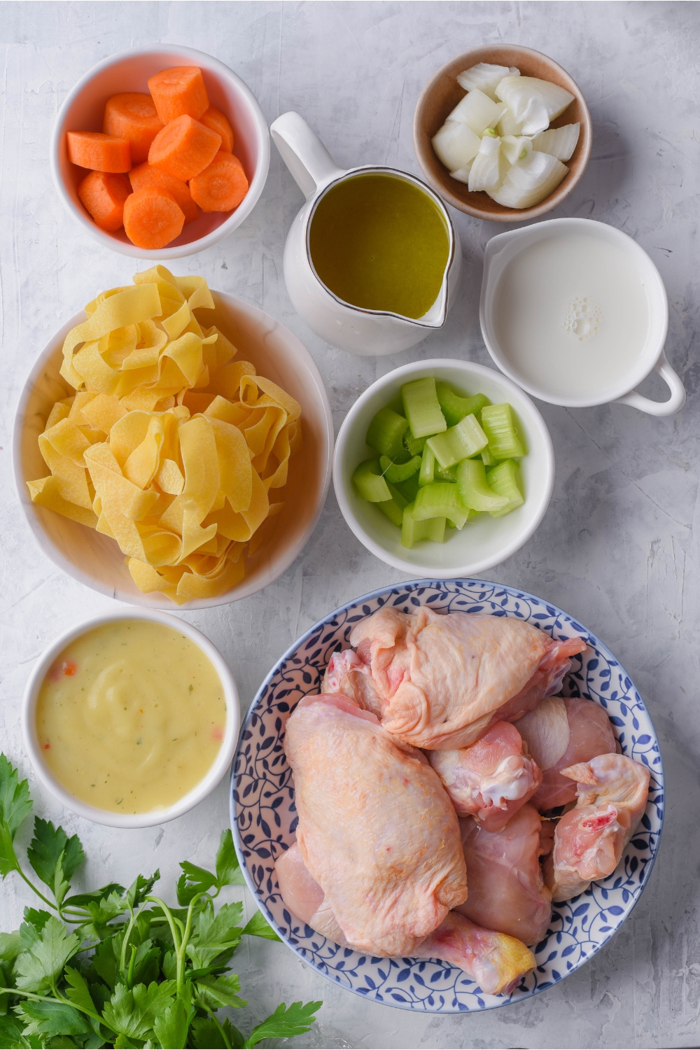 An assortment of ingredients including bowls of dried egg noodles, diced celery, carrots, onion, milk, condensed soup, and a plate of raw chicken.