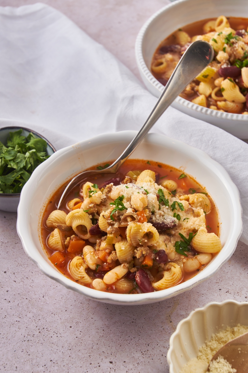 A serving of pasta e fagioli soup garnished with parmesan cheese and fresh green herbs. There is a spoon in the bowl.