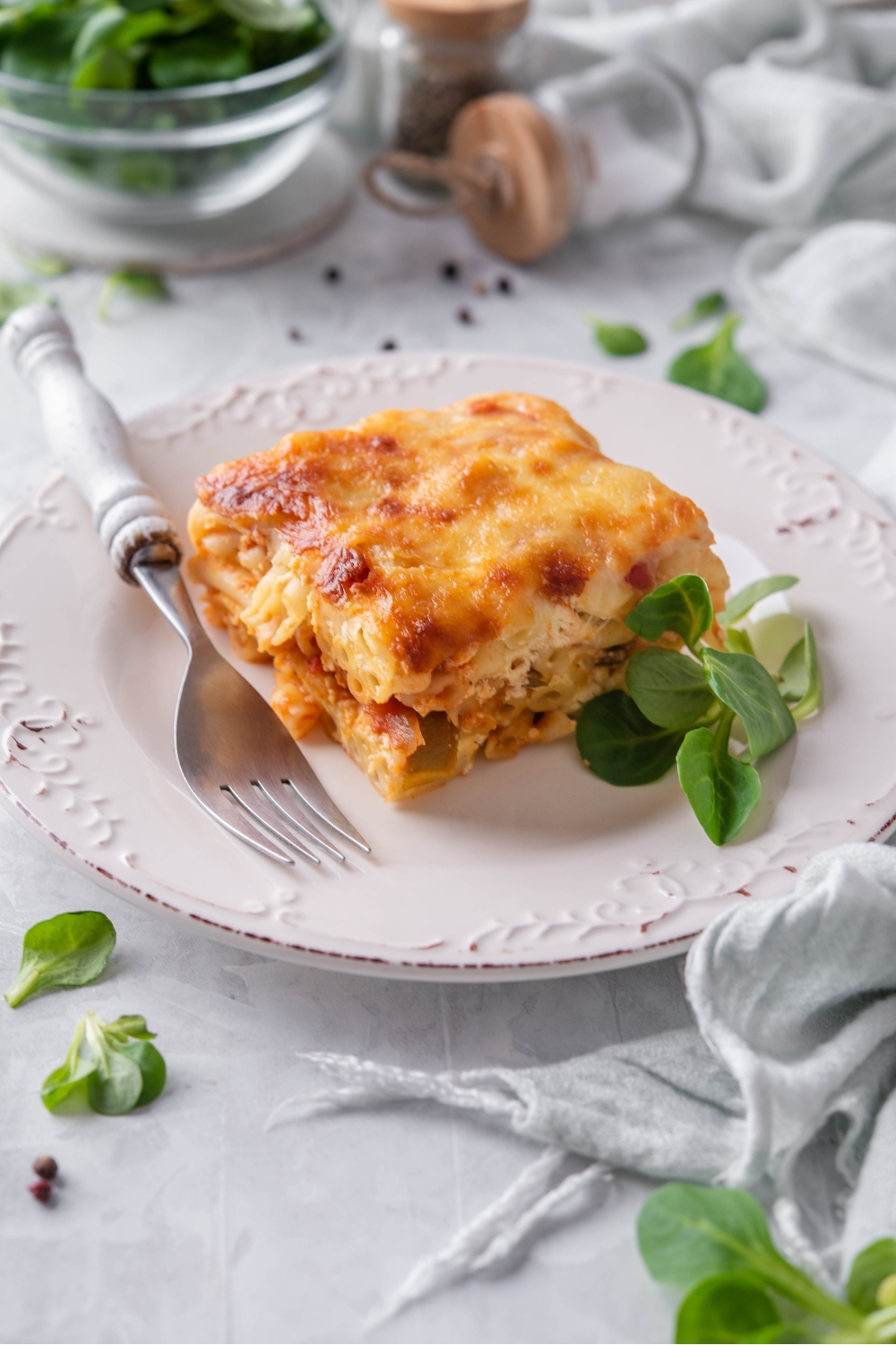 A square of baked ziti topped with golden brown cheese on a white plate with fresh greens and a fork on the plate.