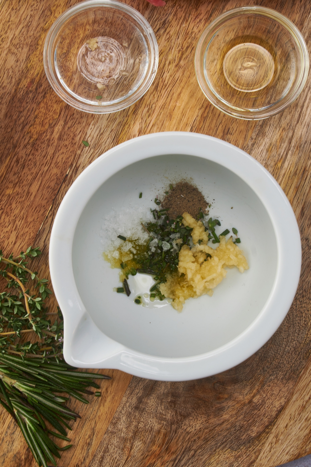 A white mortar with garlic and spices in it next to two empty bowls and fresh herbs.