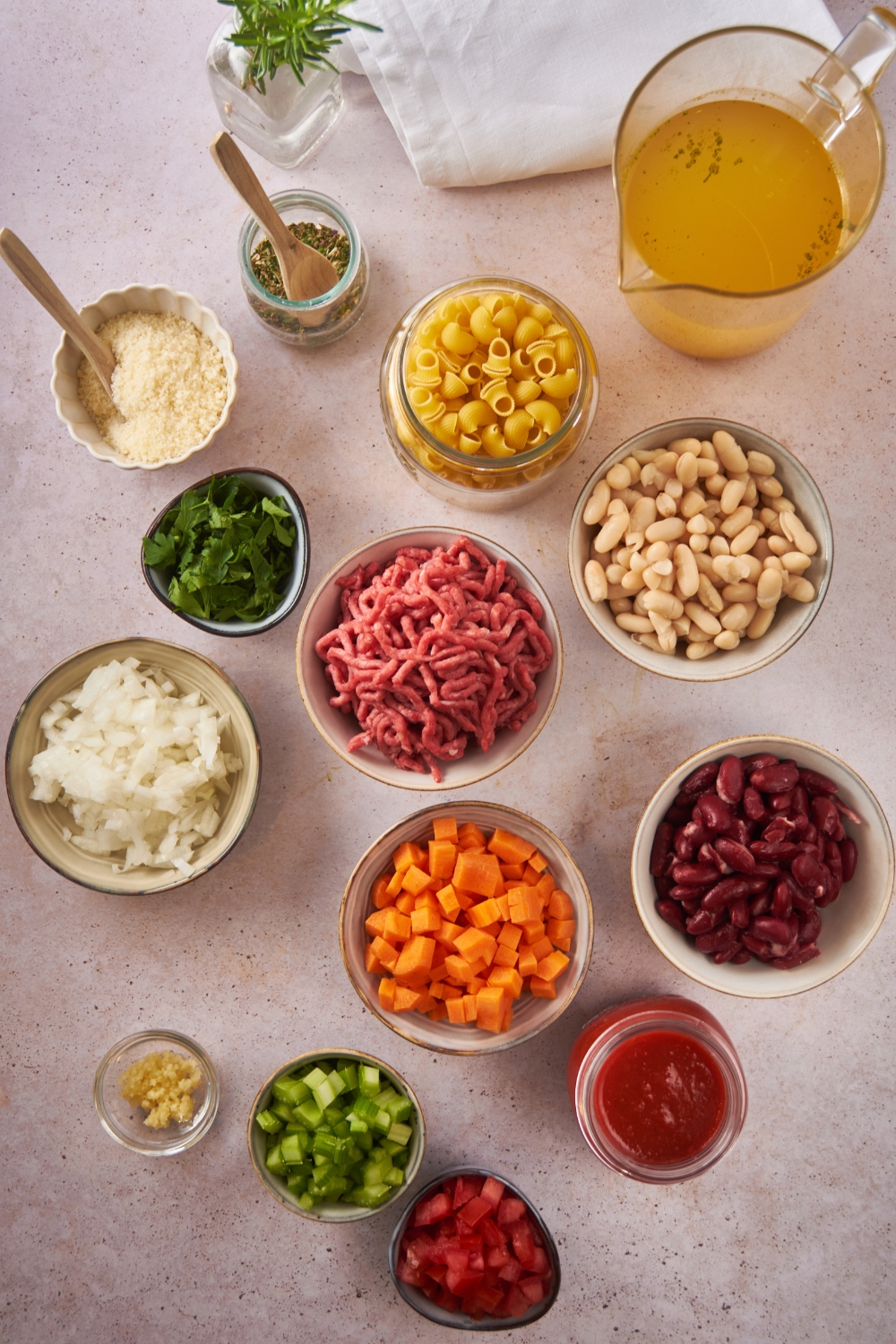 Overhead view of an assortment of ingredients including bowls of diced carrots, onion, raw ground beef, kidney beans, dried pasta, and a pitcher of chicken broth.