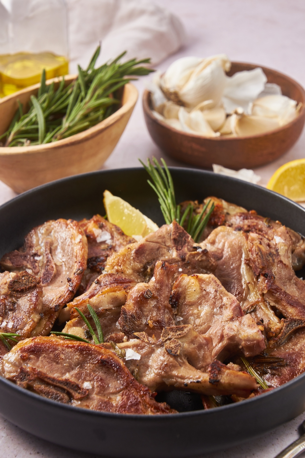 A plate of cooked baby lamb chops piled on top of each other, garnished with sea salt and fresh herbs and a lemon wedge. In the background is a bowl of rosemary and a bowl of garlic.