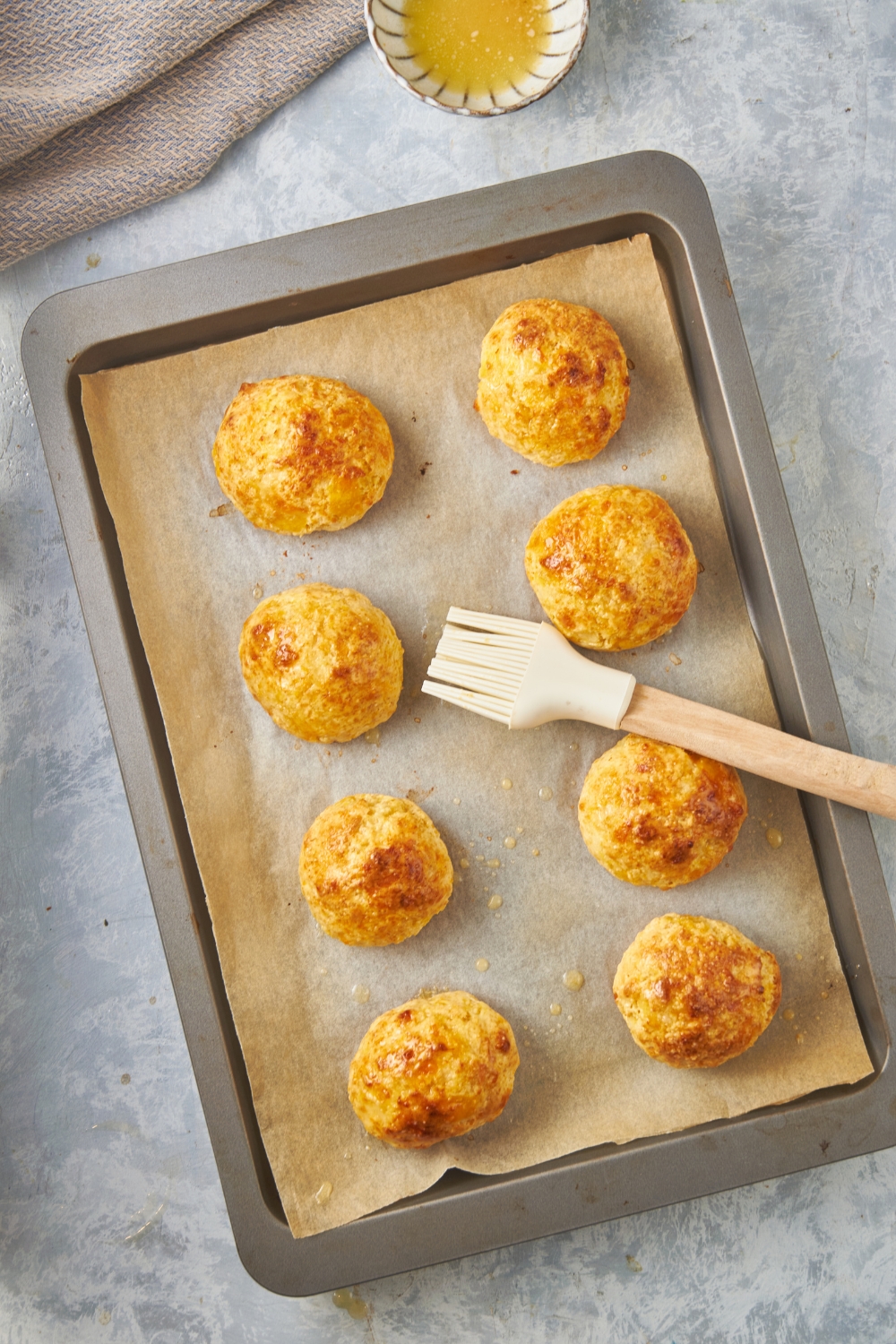 Eight equal-sized baked biscuits on a baking sheet lined with parchment paper and a silicone basting brush covered in melted butter between them.