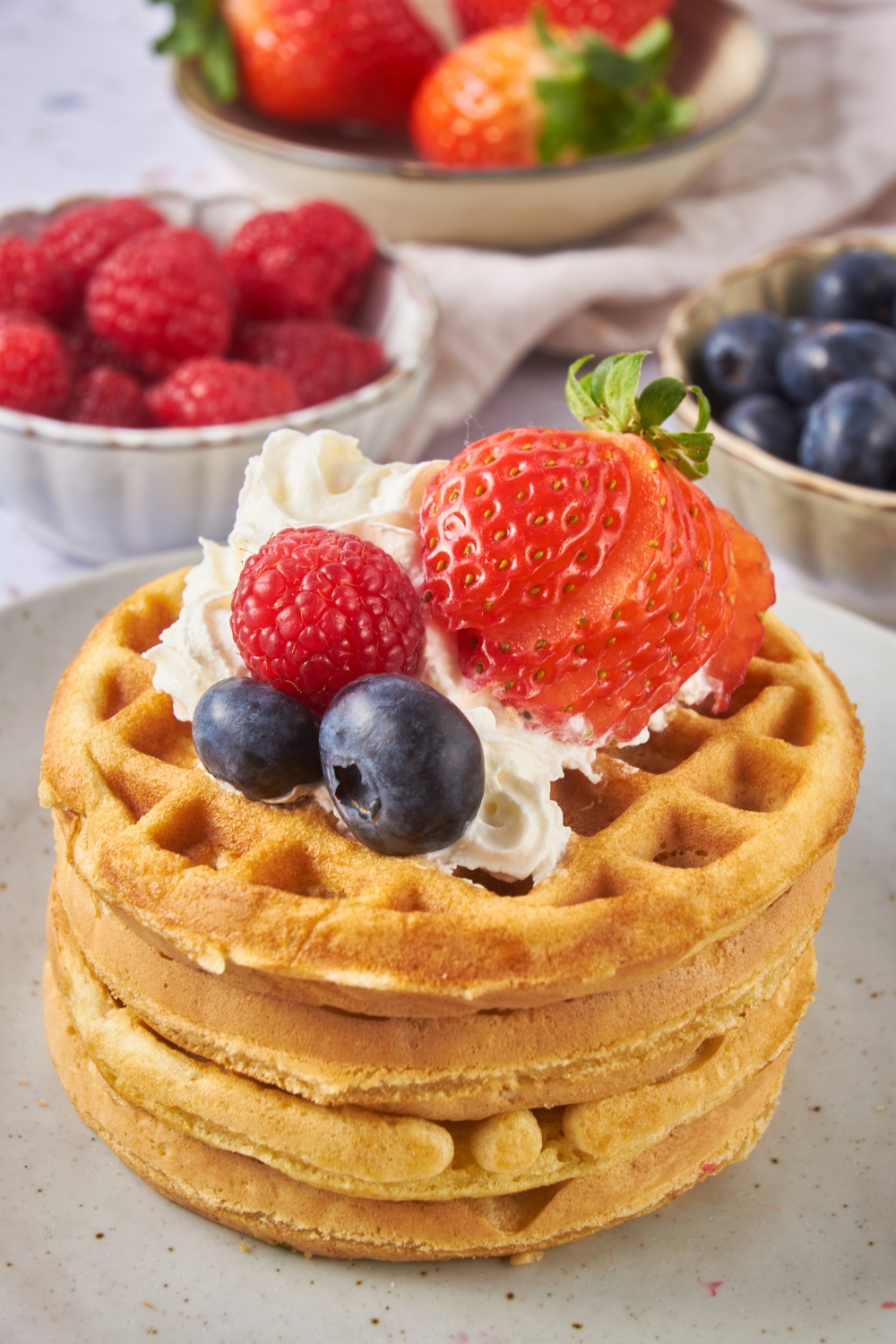 A stack of waffles topped with whipped cream and fresh berries. In the background are bowls of berries.