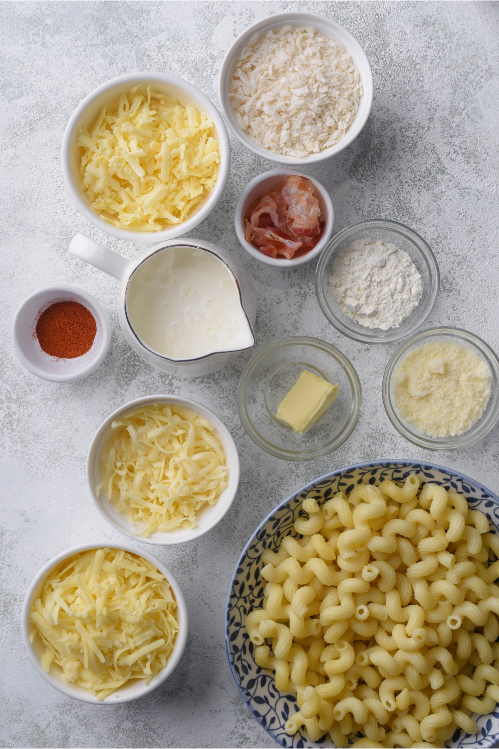 An assortment of ingredients including bowls of breadcrumbs, shredded cheese, bacon, butter, and pasta noodles.
