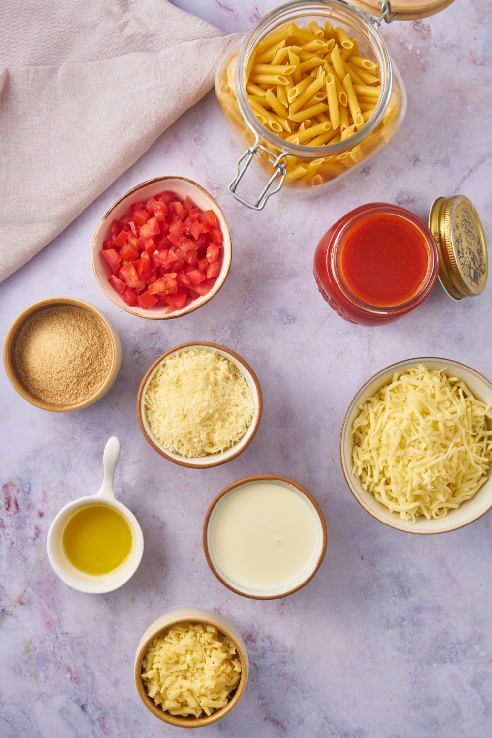 Overhead view of an assortment of ingredients including bowls of marinara sauce, diced tomatoes, dried pasta, shredded cheese, cream, and bread crumbs.