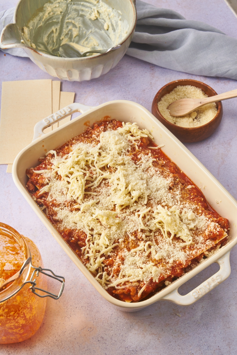 A large white baking dish with an unbaked lasagna covered in shredded cheese on top. Surrounding the lasagna are empty bowls and a bowl of parmesan cheese.