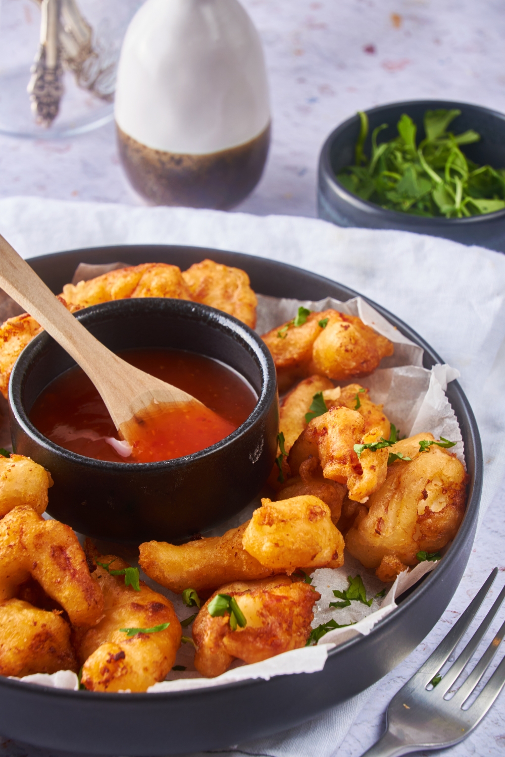 Battered and fried shrimp on a black serving plate garnished with fresh herbs and a bowl of sweet and sour sauce on the plate.