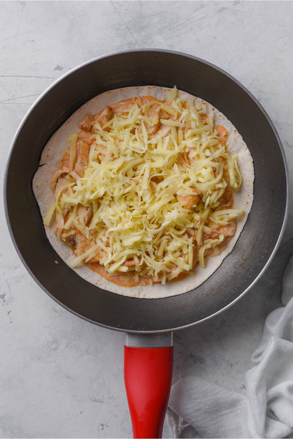 A skillet with a flour tortilla piled with chicken, sauce, and shredded cheese on top.