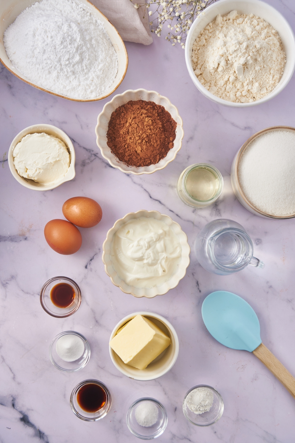 An assortment of ingredients including bowls of buttermilk, flour, cocoa powder, butter, vanilla extract, sugar, cream cheese, and two eggs.