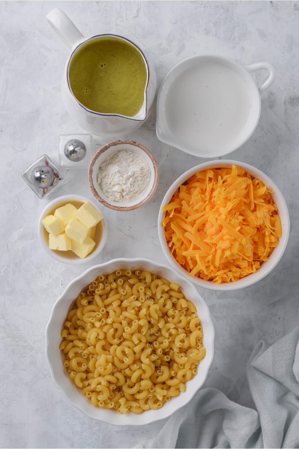 Overhead view of an assortment of ingredients including bowls of dried pasta, shredded cheese, flour, milk, butter cubes, and broth.