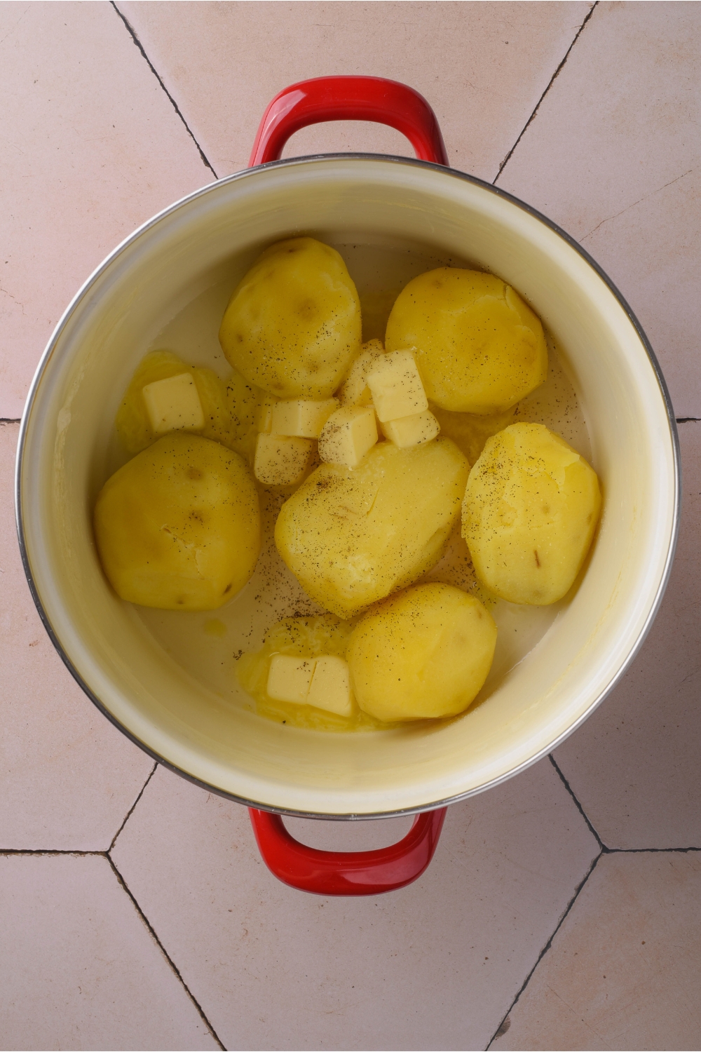 A large pot filled with peeled and boiled potatoes, butter cubes, salt, and pepper. The butter is melting in the pot.