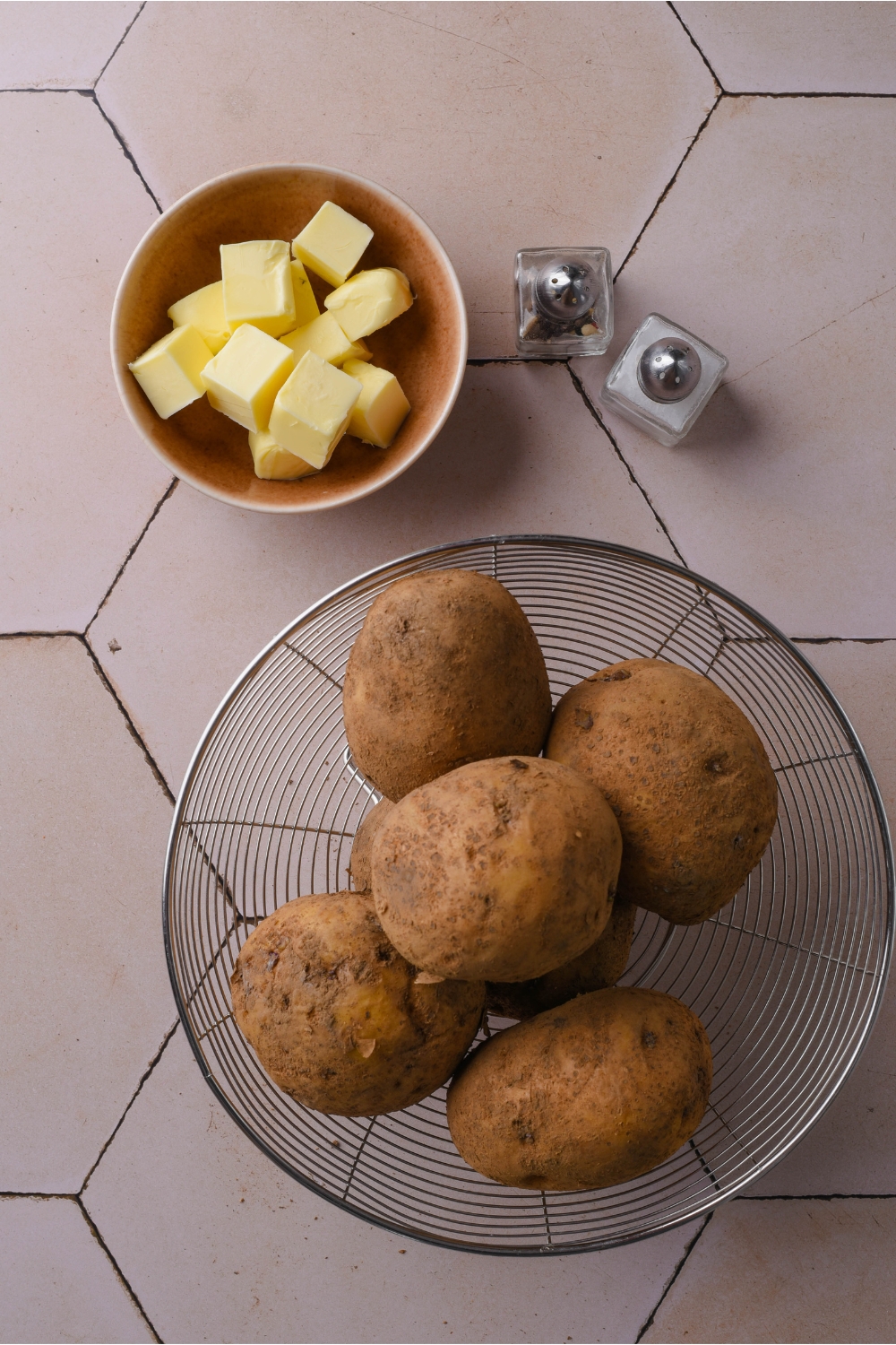 A colander filled with russet potatoes next to a bowl of butter cubes and mini salt and pepper shakers.