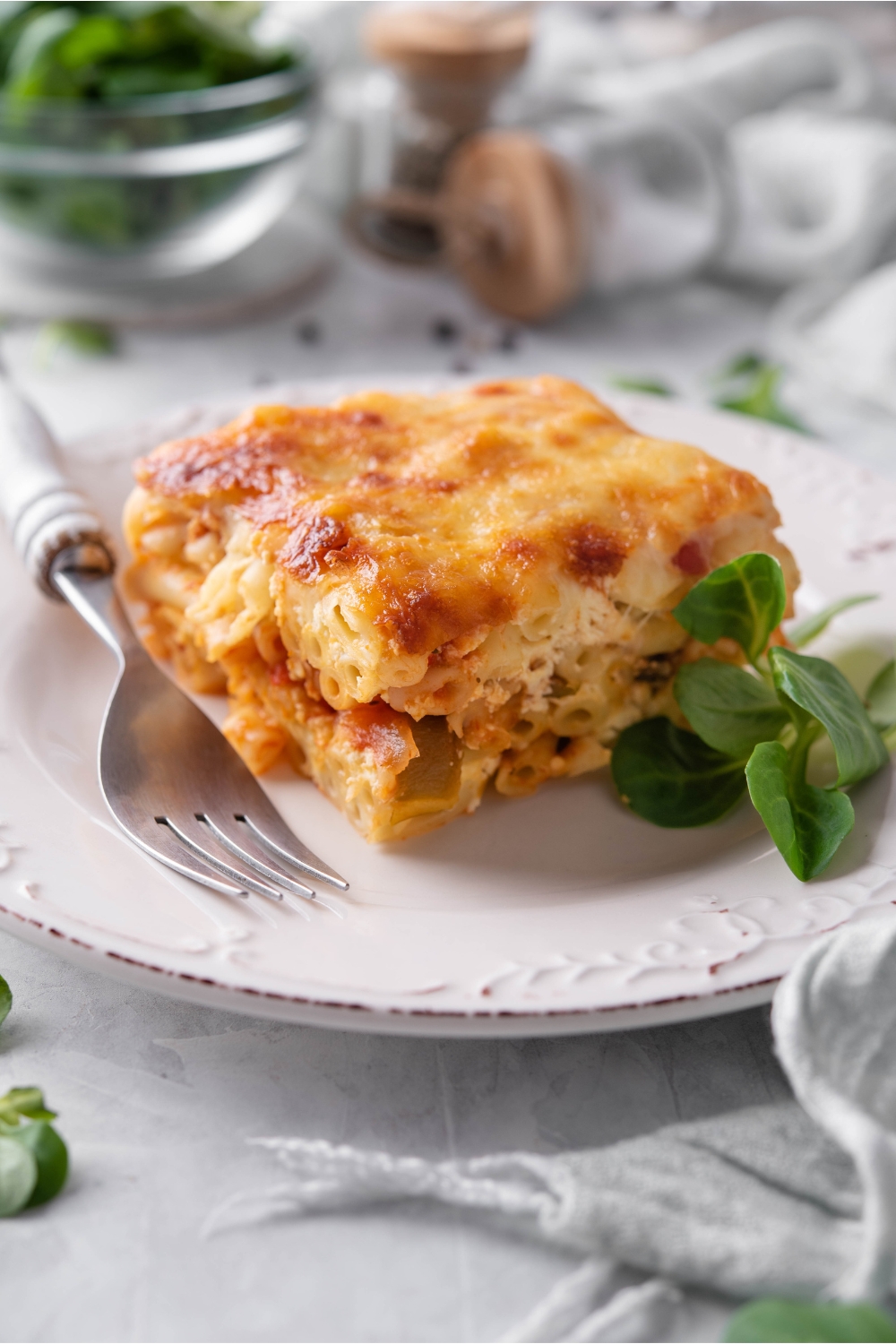 A square of baked ziti topped with golden brown cheese on a white plate with fresh greens and a fork on the plate.