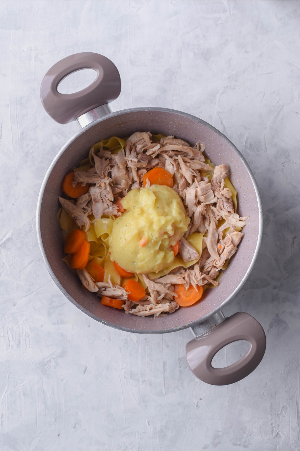 Grey pot filled with cooked shredded chicken, cooked carrots, egg noodles, and a dollop of condensed soup in the center.