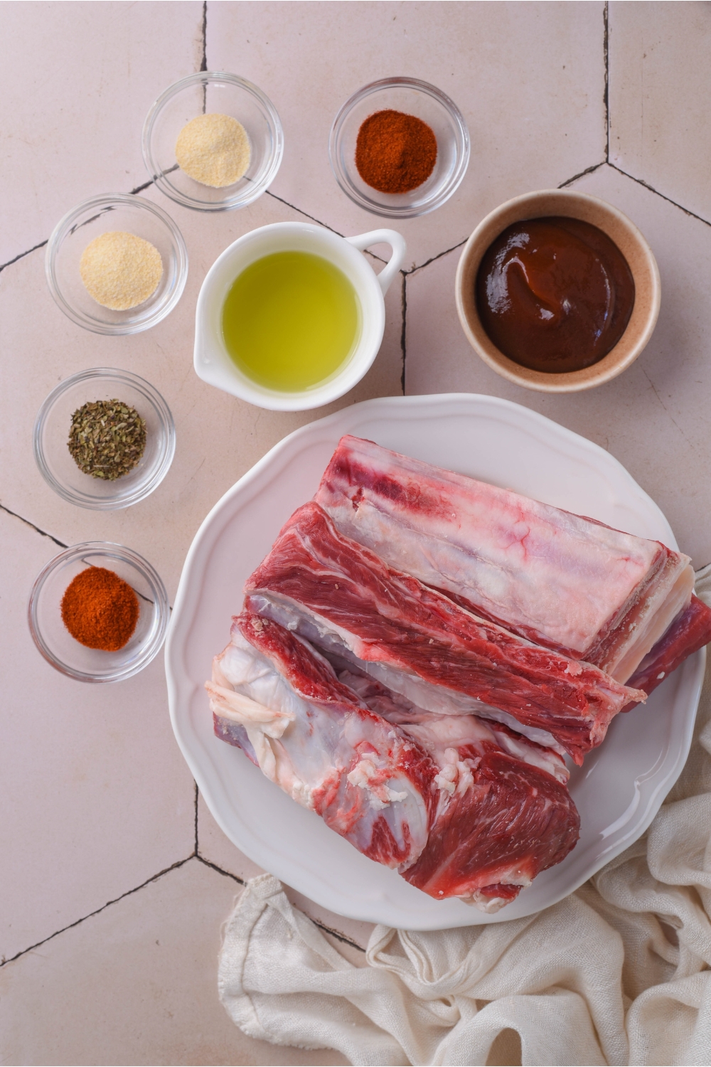An assortment of ingredients including a plate of raw beef ribs and bowls of oil, spices, and barbecue sauce.