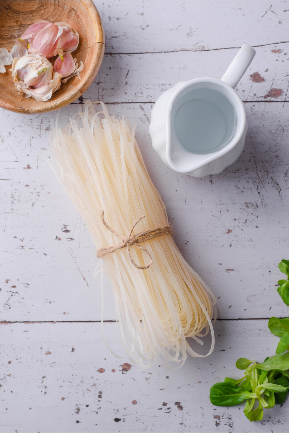 A bunch of dried rice noodles tied together by twine next to a pitcher of water and a bowl of garlic.