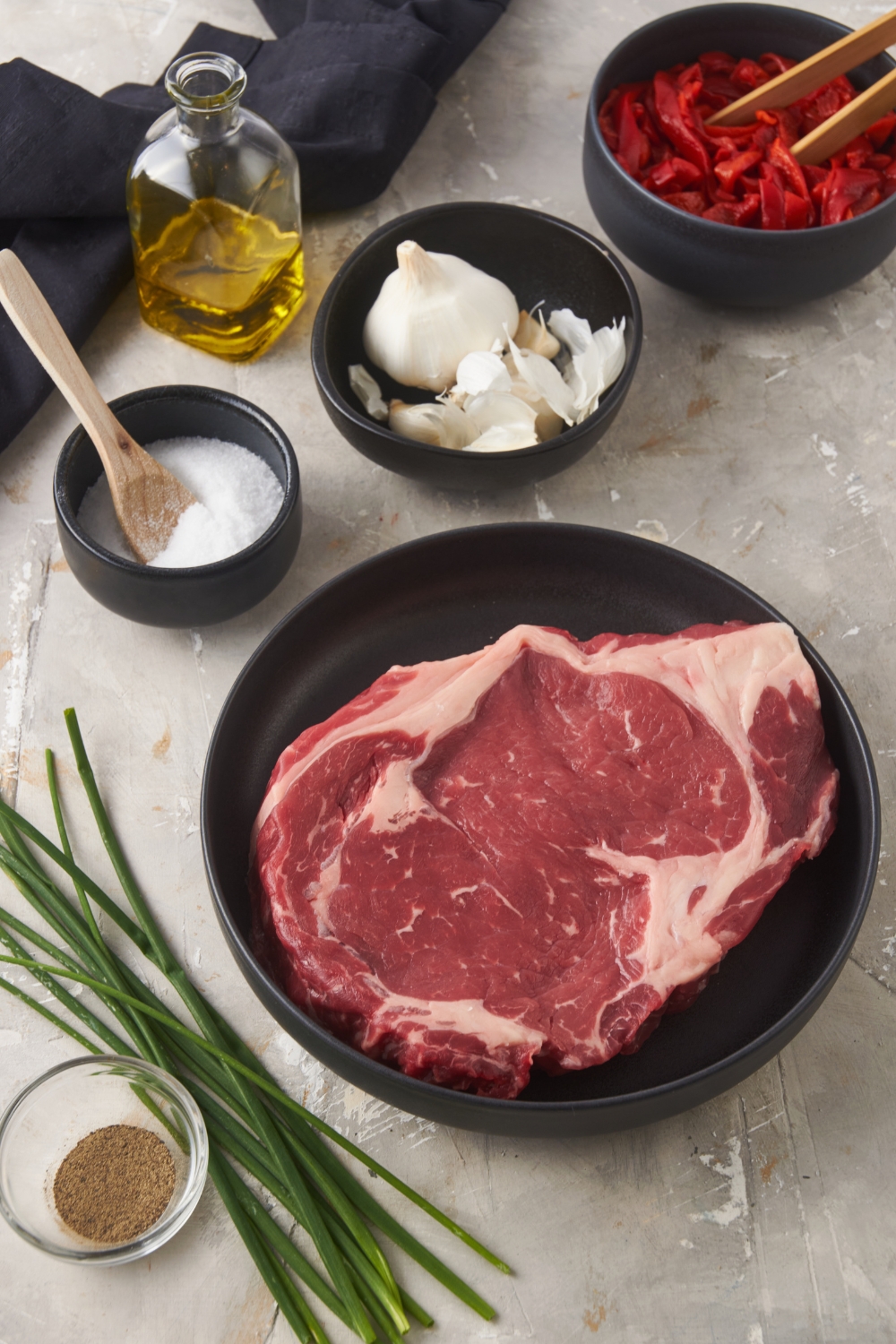 An assortment of ingredients including a plate of ribeye steak, a bowl of garlic, a bowl of salt, pepper, and a bunch of chives.