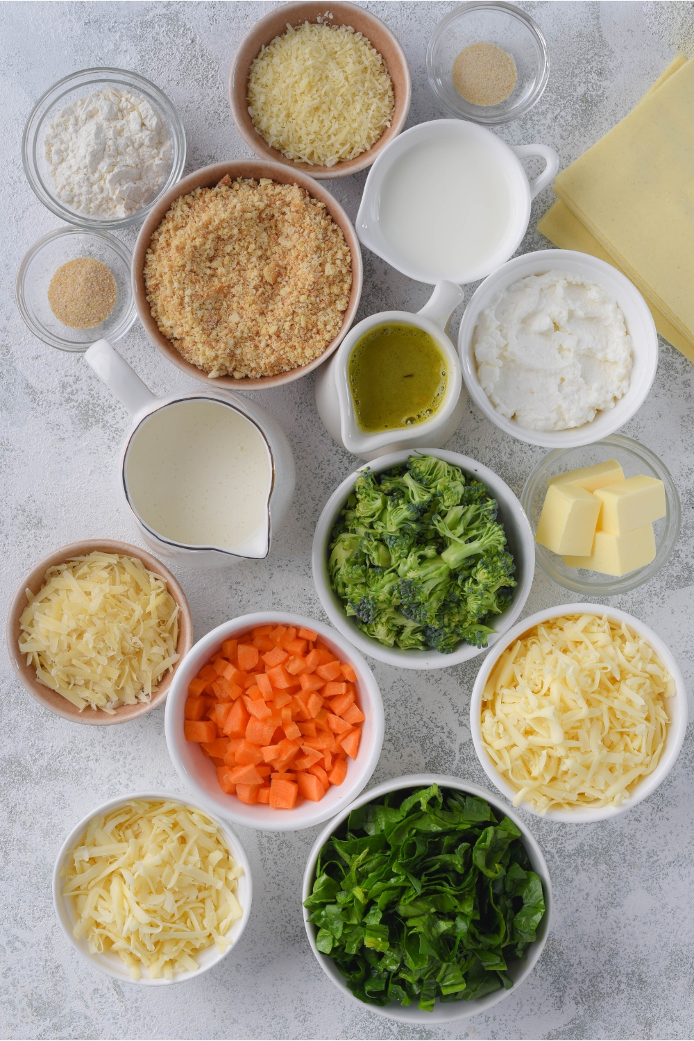 Overhead view of an assortment of ingredients including bowls of cream, ricotta cheese, cracker crumbs, diced broccoli, carrots, fresh spinach, shredded cheeses, spices, and a stack of lasagna noodles.