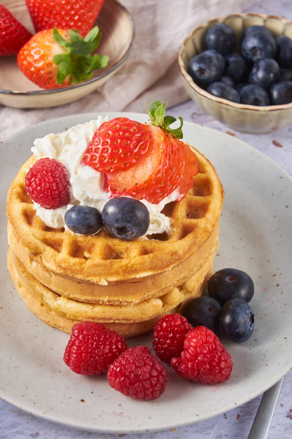 A stack of waffles topped with whipped cream and fresh berries, with some berries spilled out on the plate next to the waffles.