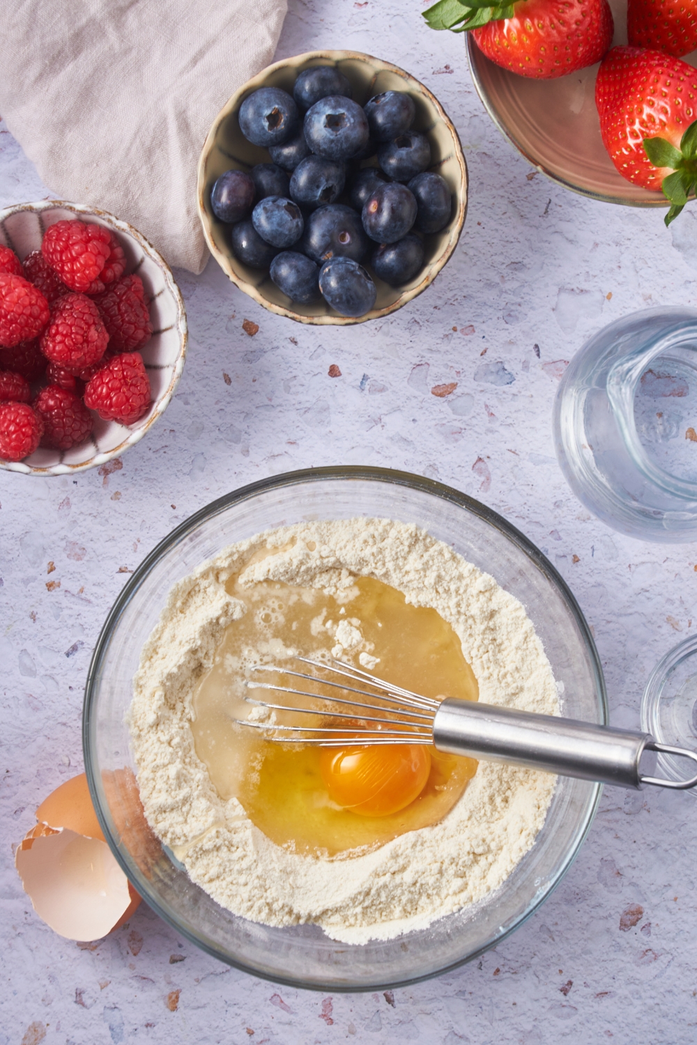 A clear bowl with waffle mix, an egg, and a whisk in it. The bowl is next to small bowls of berries.