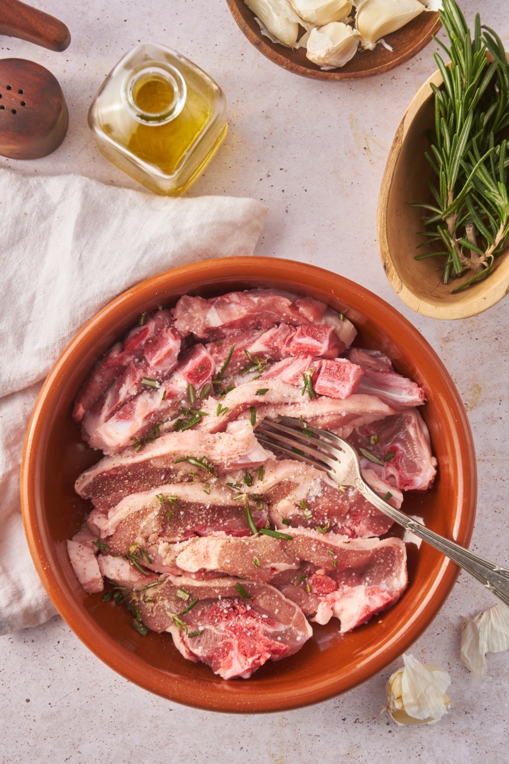 A red bowl filled with raw, seasoned lamb chops surrounded by an assortment of ingredients including a bottle of oil, garlic, and rosemary.