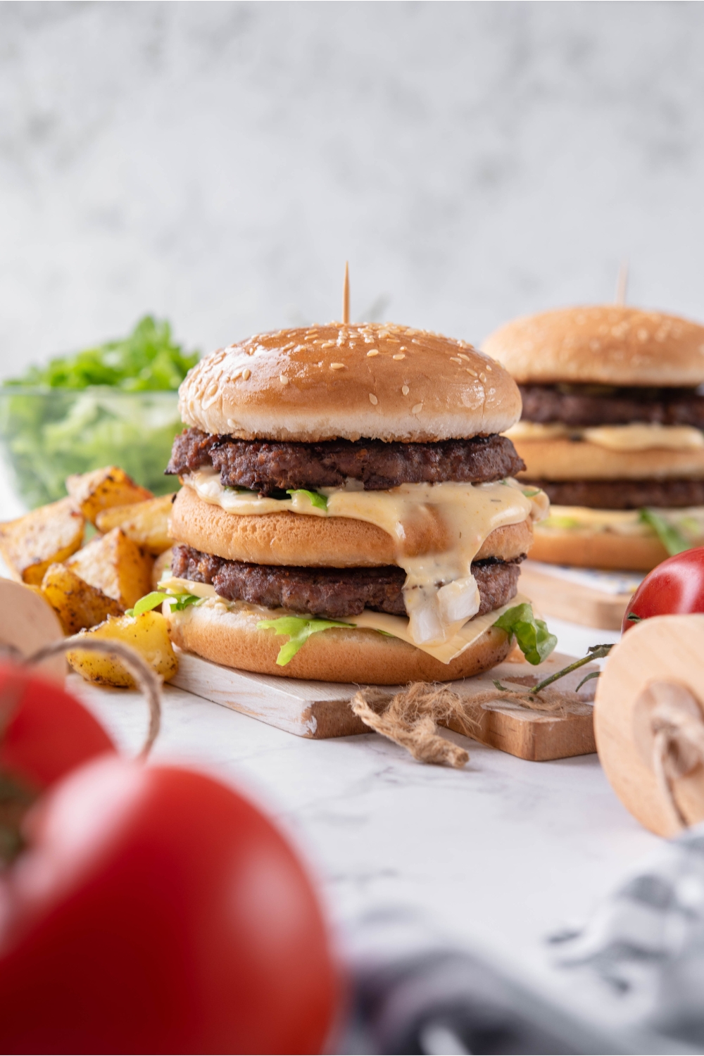 Close up of a McDonald's big mac on a wooden board with big mac sauce dripping off the side and a toothpick stuck in the center of the burger. The burger is surrounded by potato wedges and there's a second burger in the background.