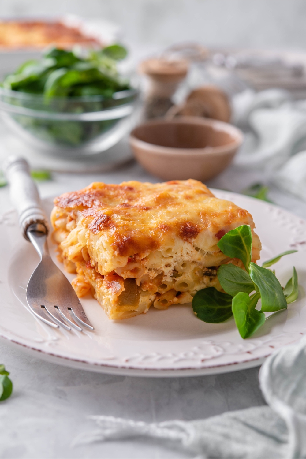 A square of baked ziti topped with golden brown cheese on a white plate with fresh greens and a fork on the plate. There is a bowl of greens in the background.