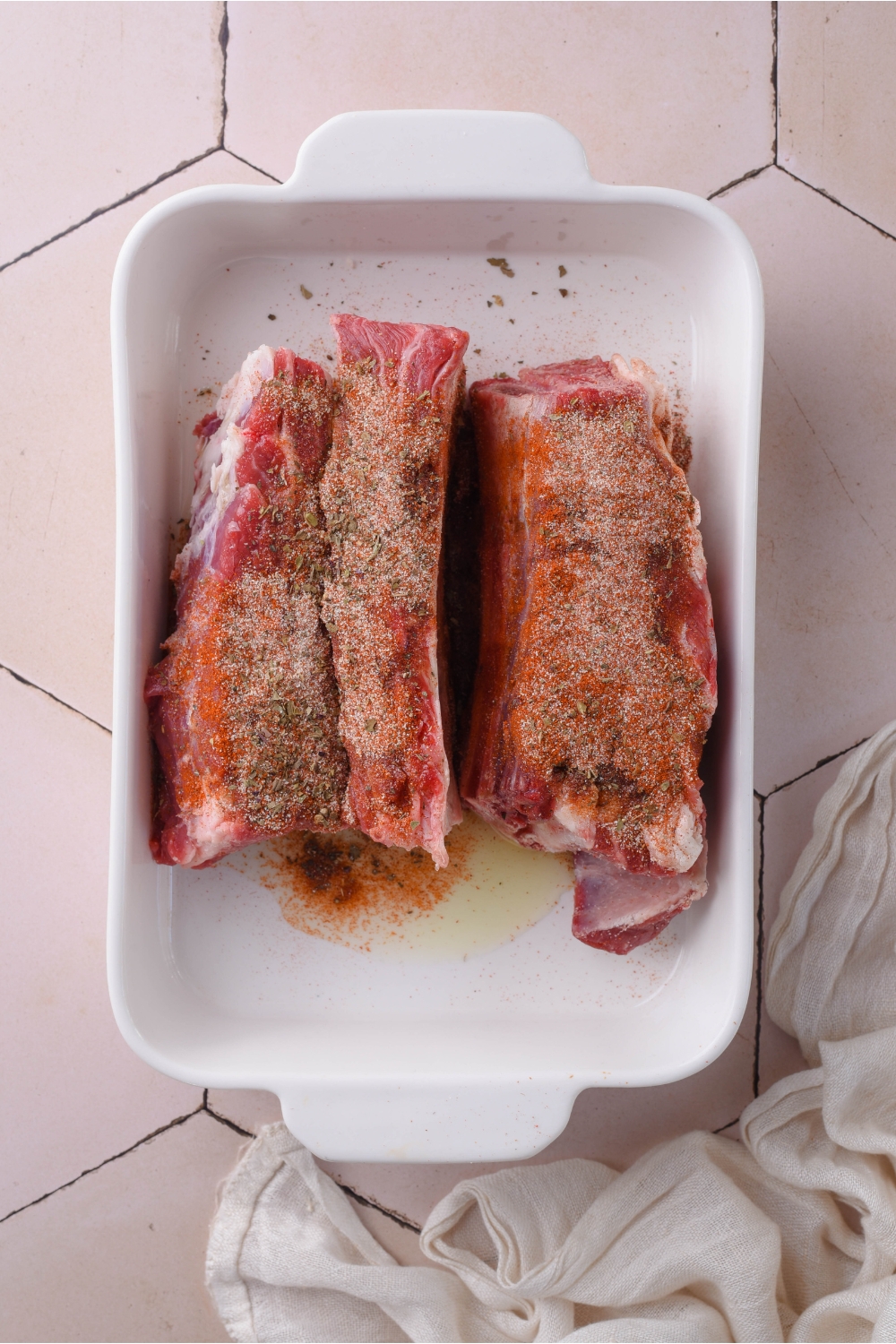 A white baking dish filled with uncooked beef ribs covered in oil and a seasoning blend.