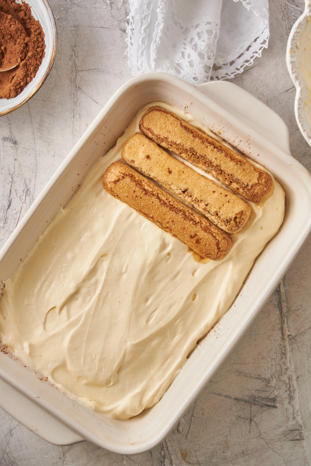 A pan with a layer of ladyfingers and custard; another layer of ladyfingers is being added.