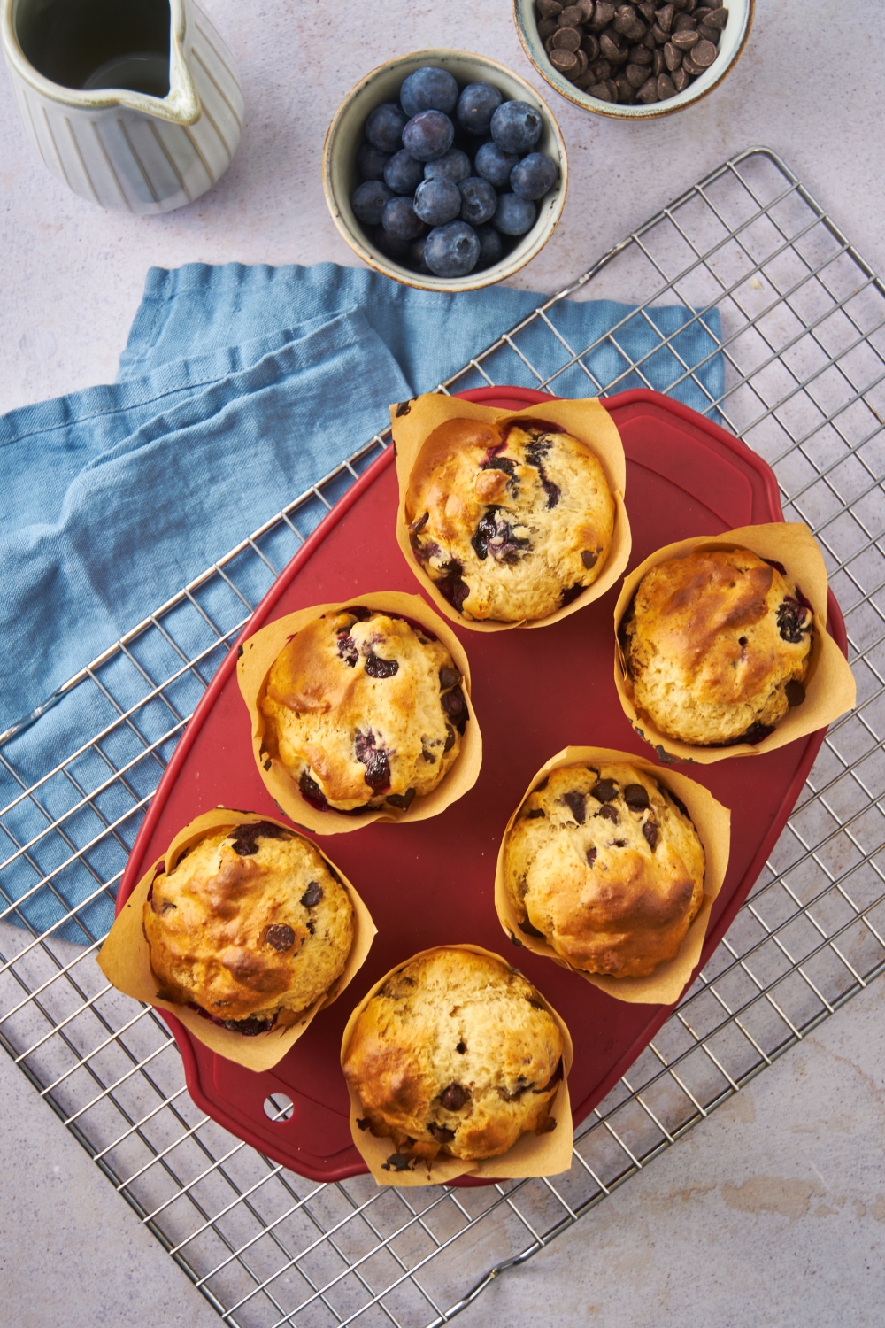 Six freshly baked chocolate chip and blueberry muffins on a red tray atop a wire rack.