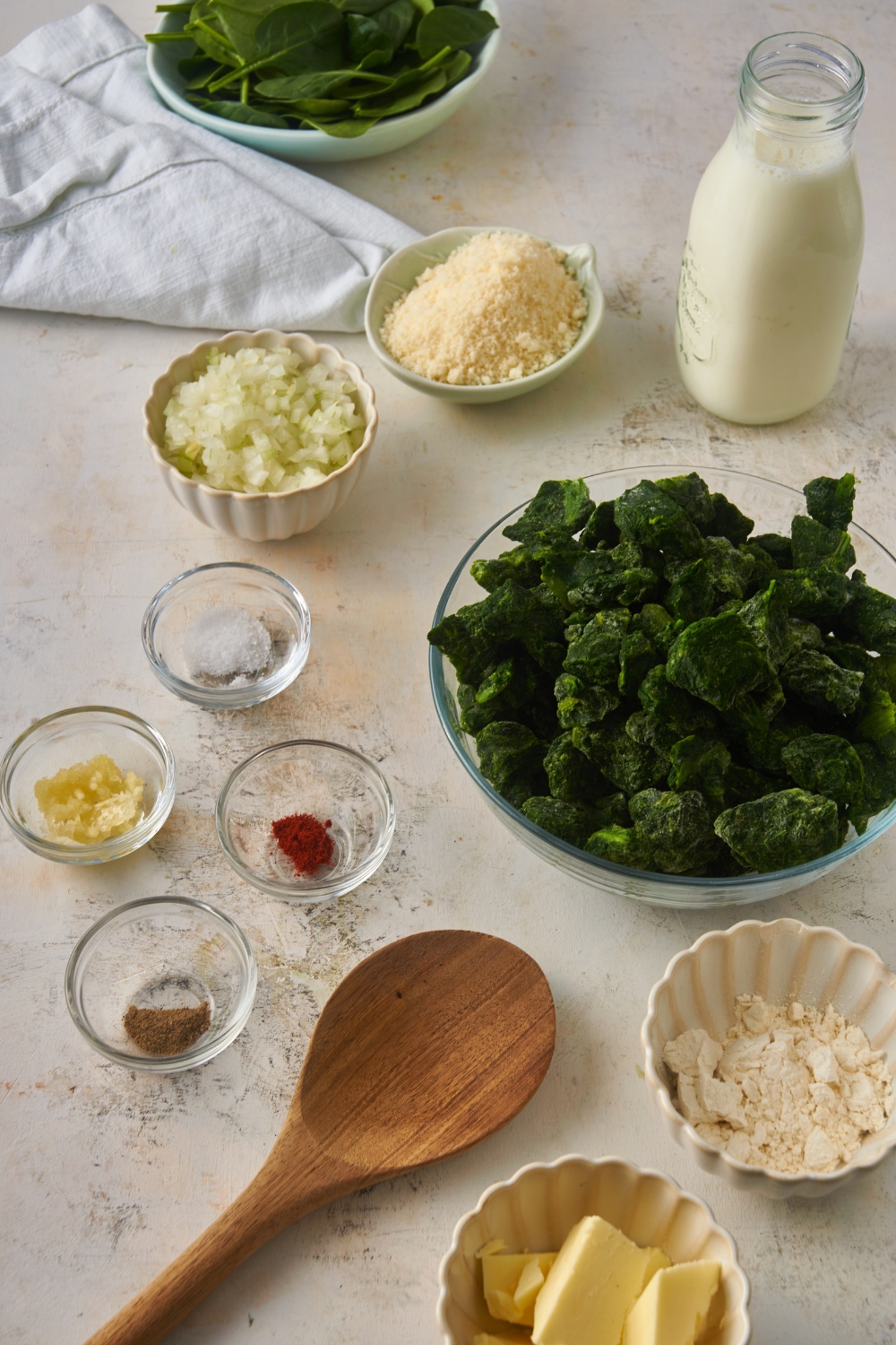 An assortment of ingredients including a bowl of frozen spinach, minced garlic, spices, diced onion, parmesan cheese, and a jar of milk.