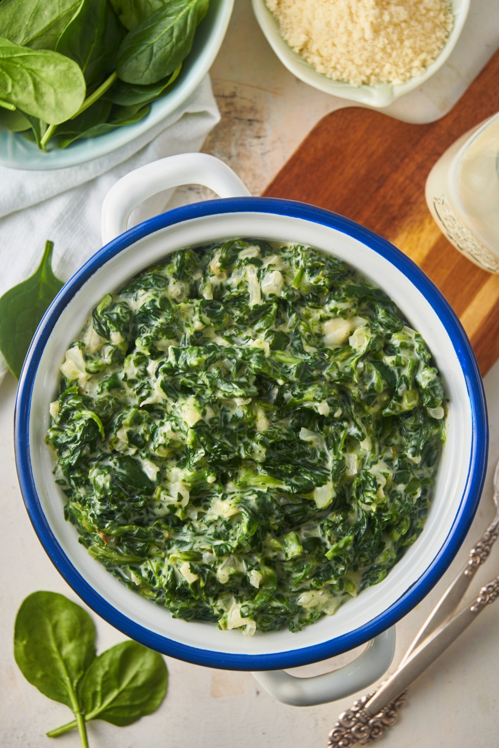 Overhead view of creamed spinach in a white and blue serving bowl atop a wooden board with fresh spinach sprinkled around the serving dish.