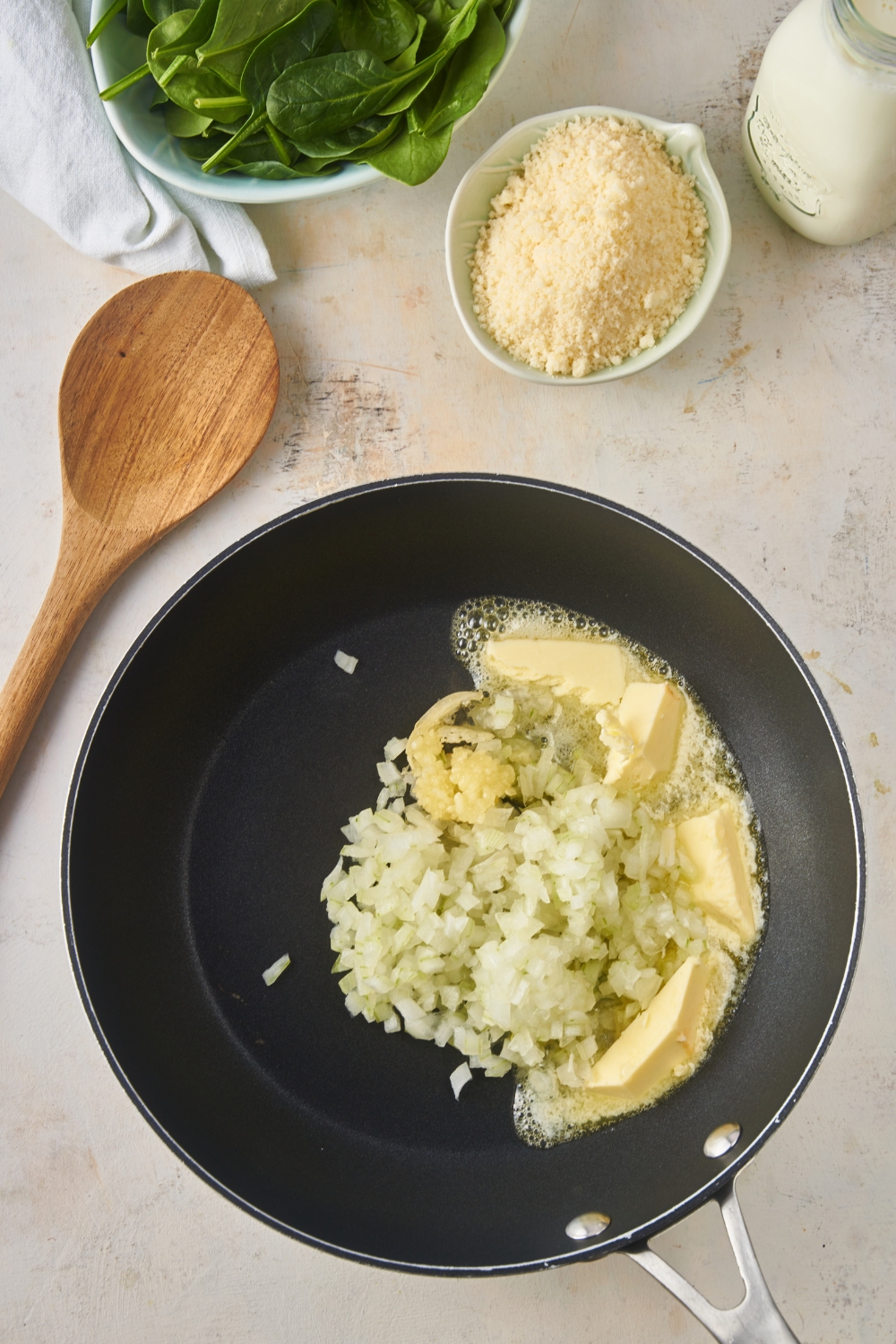 Black skillet with butter melting in it and a pile of diced onions. Next to the skillet is a wooden spoon, a bowl of parmesan cheese, and a bowl of spinach.