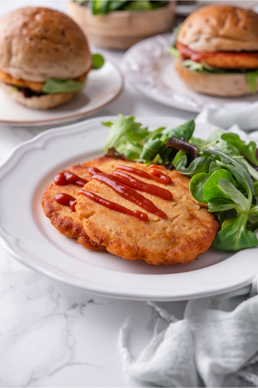 Two fried salmon patties covered with a drizzle of ketchup and layered on top of each other on a white plate with a side salad. In the background are two salmon burgers.
