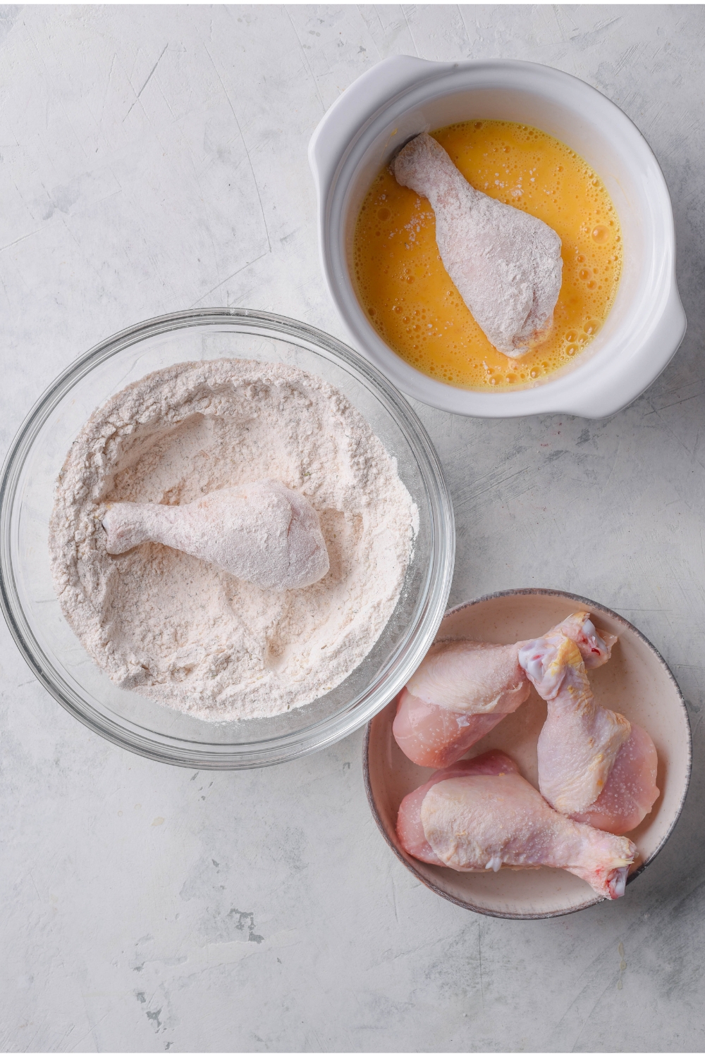 Three bowls of raw chicken, a dry flour mixture with a piece of chicken in it, and a bowl of beaten eggs with a piece of flour-coated chicken in it.