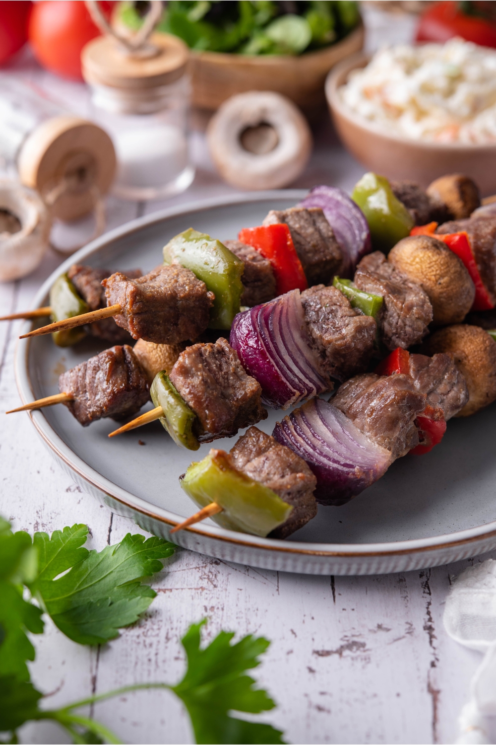 A stack of five baked steak kebabs filled with seasoned steak, bell peppers, mushrooms, and red onions. In the background are bowls of coleslaw and salad.
