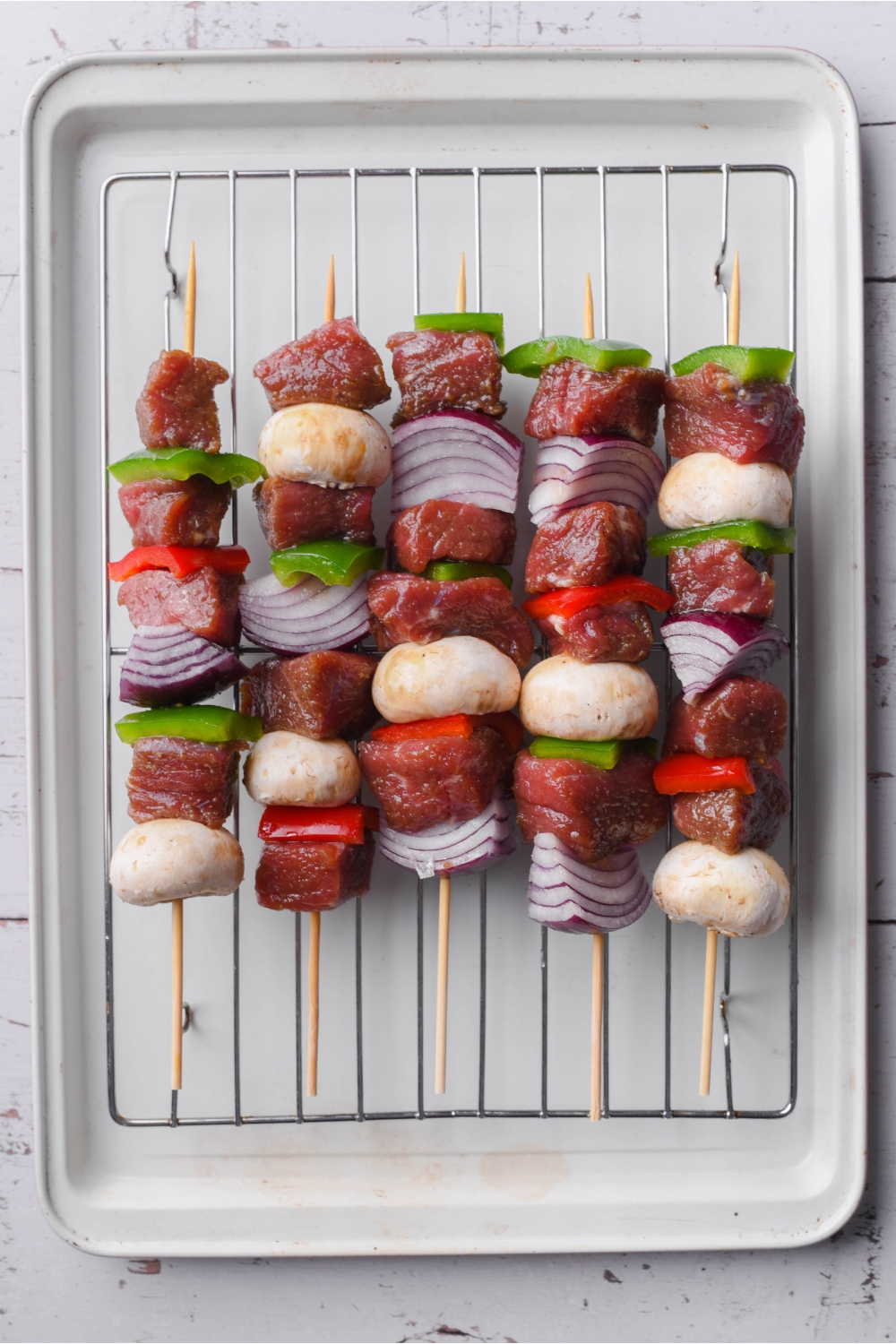 Five uncooked skewers of marinated steak, bell peppers, red onions, and mushrooms lined on a wire rack atop a baking sheet.